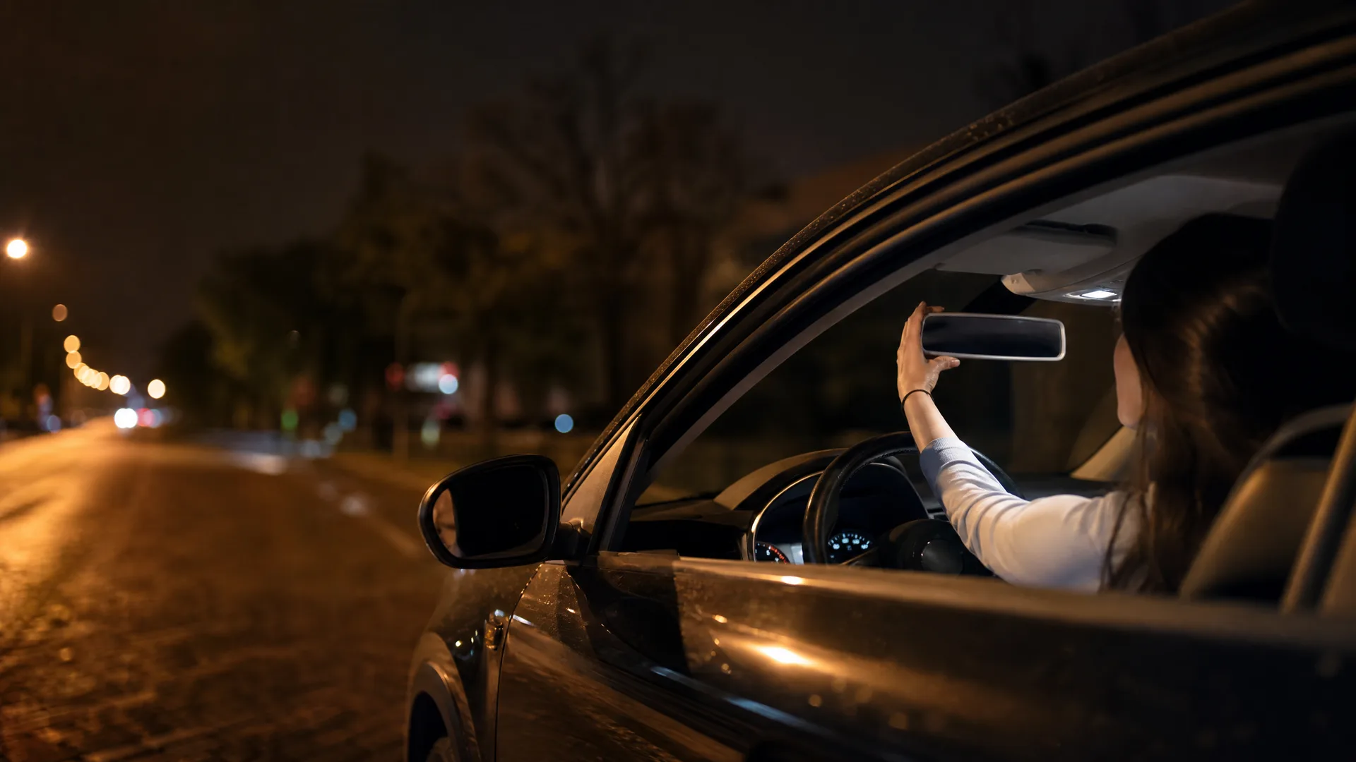 At night, a woman seated in the driver's seat of a car reaches up to adjust the rear-view mirror, seen through the open side window, with a dark street and blurred lights behind.